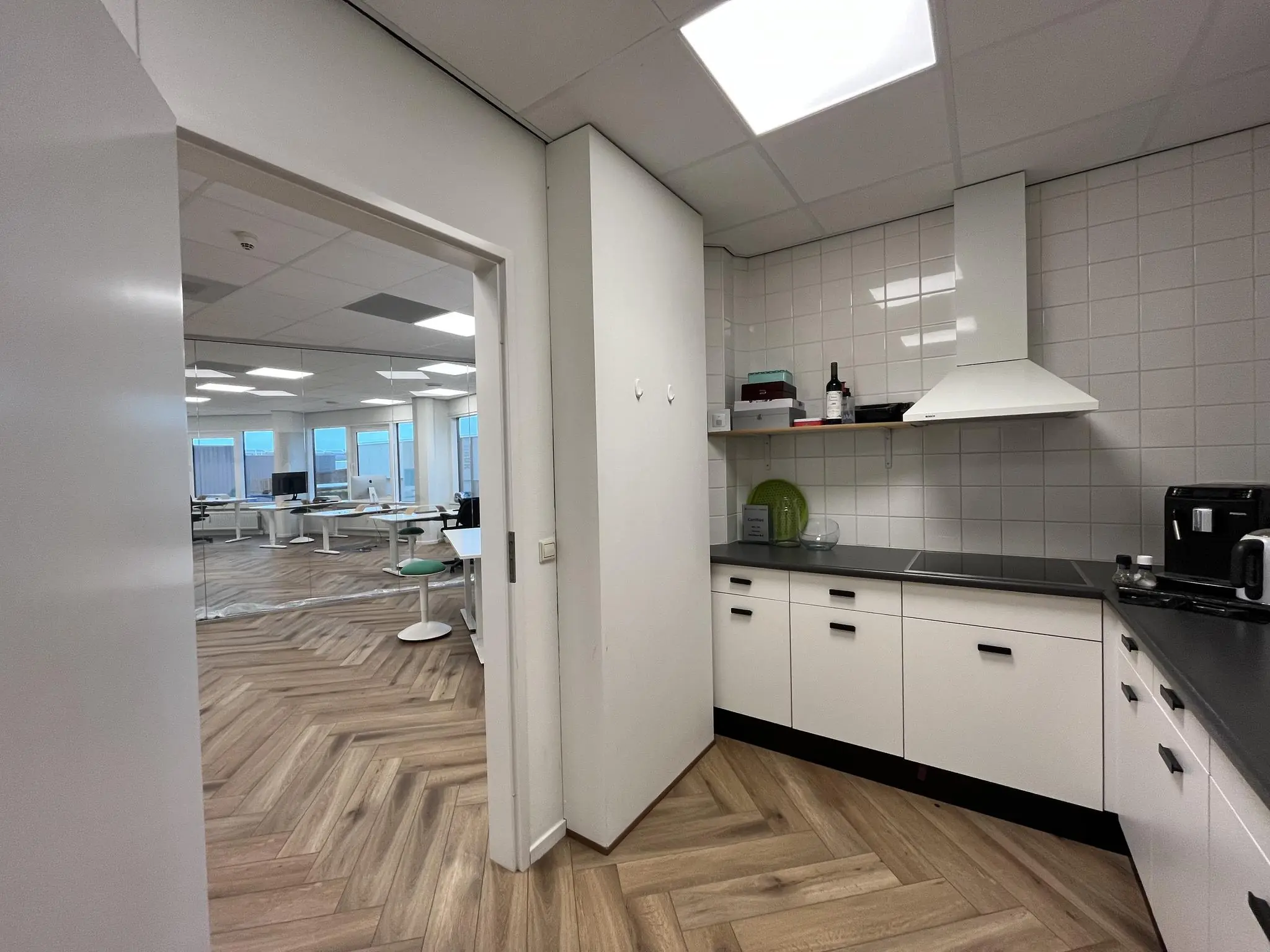 Moderne kantoorkeuken met witte kasten, zwarte accenten en zicht op een open kantoorruimte aan de Lange Dreef.